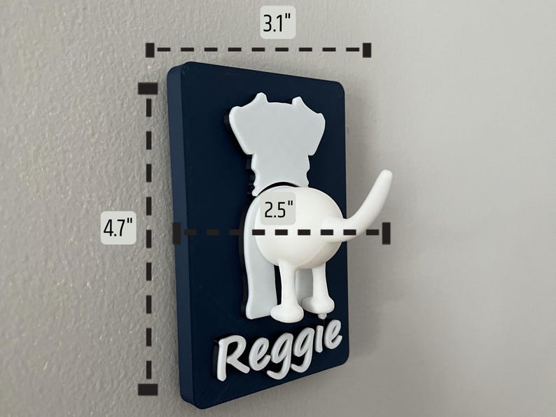 May include: A white dog-shaped hook with a blue background and the name "Reggie" printed on it. The hook measures 4.7 inches tall and 3.1 inches wide.