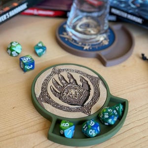 Puede incluir: Una bandeja de dados verde y marrón con un diseño de pata de oso. La bandeja tiene cuatro ranuras para dados y un área más grande para tirar. Hay varios dados multicolores en la bandeja.