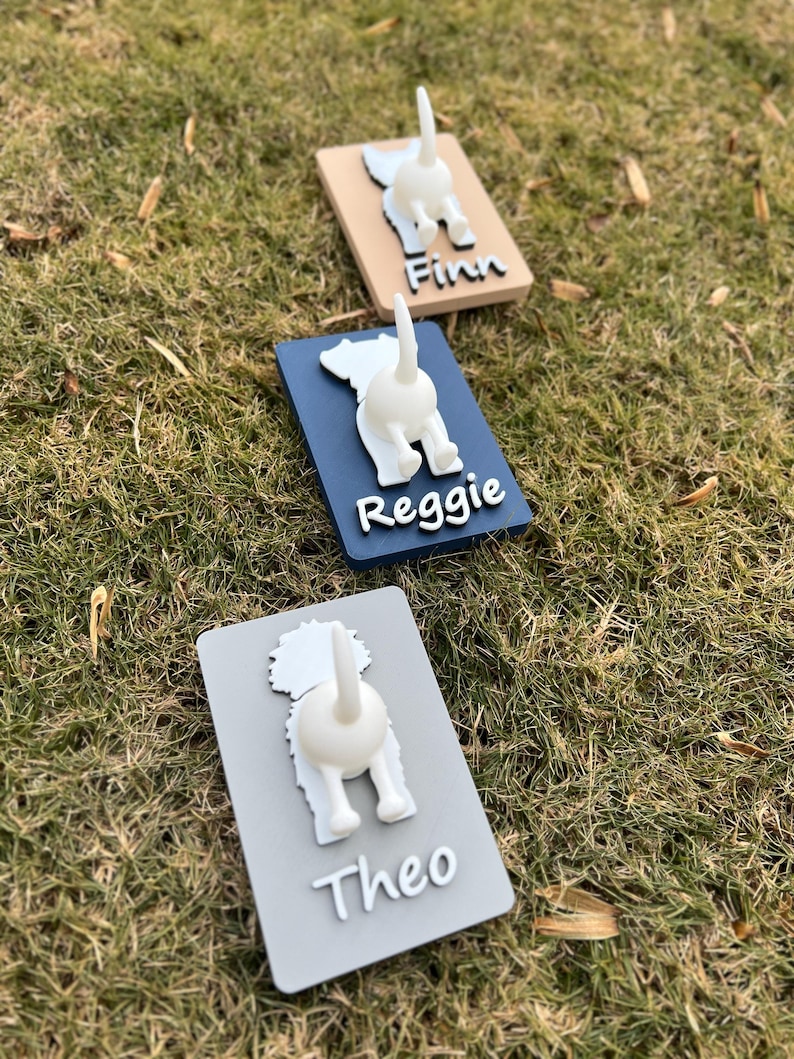 May include: Three dog-shaped hooks with the names "Finn", "Reggie", and "Theo" written on them. The hooks are made of wood and painted in different colors: beige, blue, and gray.