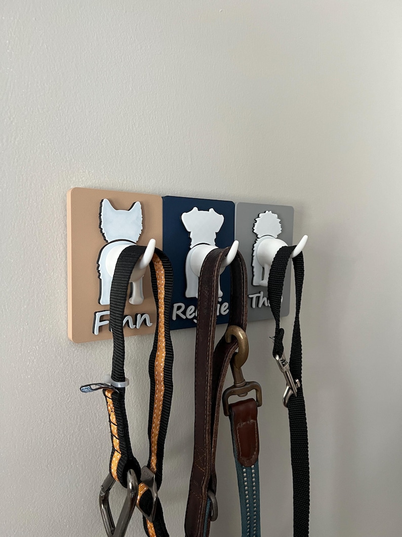 May include: Three dog-shaped hooks with leashes hanging from them. The hooks are painted in different colors: beige, blue, and gray. The leashes are black, brown, and blue. The hooks are labeled "Finn", "Reggie", and "Theo".