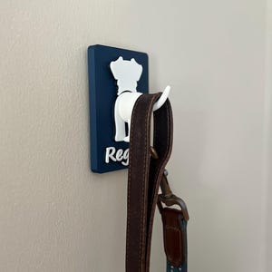 May include: A blue and white dog-shaped hook with the name "Reg" on it. A brown leather leash is hanging from the hook.