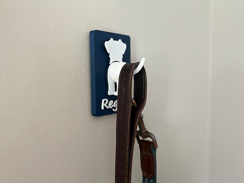 May include: A blue and white dog-shaped hook with the name "Reg" on it. A brown leather leash is hanging from the hook.