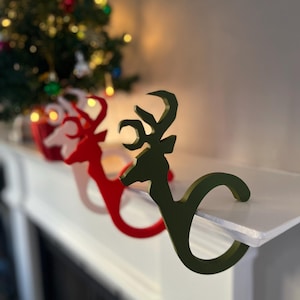 May include: Three Christmas stocking holders shaped like a deer head. The holders are in red, white, and green. The green holder is in the foreground and the red and white holders are in the background.