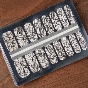 May include: A sheet of nail polish strips with a white base and a black scribble design. The nail strips are in a clear plastic package, and the background is a dark brown wooden surface.