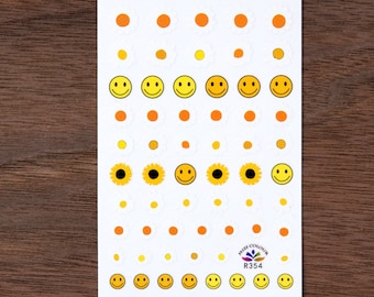 May include: A sheet of yellow and orange nail stickers with smiley faces and sunflowers. The stickers are arranged in rows and are ready to be applied to nails.