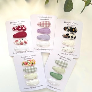 May include: Five sets of hair clips on white cards. Each set has three clips with different patterned fabrics. The fabrics include gingham, floral, polka dots, and animal print. The cards have the text "Thoughts of Grace Accessories" and a website address.