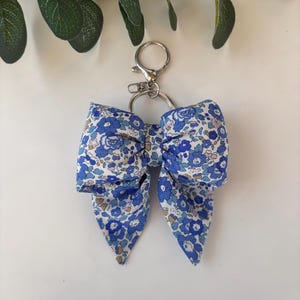 May include: A fabric bow keyring featuring a blue and white floral pattern. The bow is attached to a silver-toned metal split ring and a lobster claw clasp. The fabric has a repeating print of small blue flowers with light brown accents.