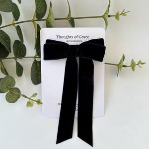 May include: A black velvet bow hair clip with long, flowing tails. The bow is centered on a white card with the text "Thoughts of Grace Accessories". The background includes green leafy sprigs.