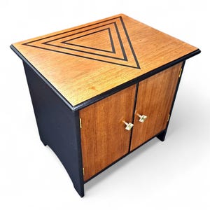 May include: A small wooden cabinet with a geometric design. The top is light brown with a black triangle design. The cabinet has two doors with gold-coloured knobs and a dark blue base.