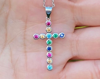 Puede incluir: Un collar de cadena de plata con un pequeño colgante en forma de cruz. La cruz está adornada con piedras preciosas de colores, incluyendo rosa, azul, verde y amarillo.