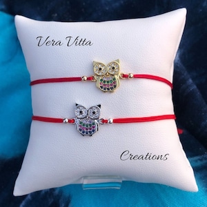 May include: Two owl charm bracelets on a white pillow. One bracelet has a gold owl charm, the other has a silver owl charm. Both have red cords. The text "Vera Vitta" and "Creations" are visible.