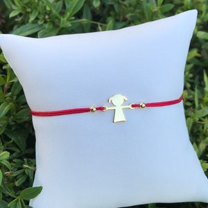 Puede incluir: Una pulsera de cordón rojo con un dije dorado en forma de persona. La pulsera también presenta dos pequeñas cuentas doradas. La pulsera se exhibe sobre una almohada blanca sobre un fondo de follaje verde.