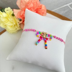 Op de afbeelding: Een roze kralen armband met een kleurrijke strik bedel. De strik is gemaakt van kleine, kleurrijke kralen en heeft een gouden accent in het midden.