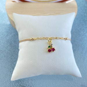 Op de afbeelding: Een gouden kettingarmband met een rode en groene kers-hanger. De hanger is versierd met kleine, heldere kristallen.