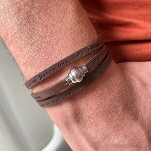 May include: A brown leather bracelet with a silver clasp. The bracelet has the words "as we forgive those who trespass against us" and "Our Father, who art in heaven, hallowed be thy name" engraved on it.