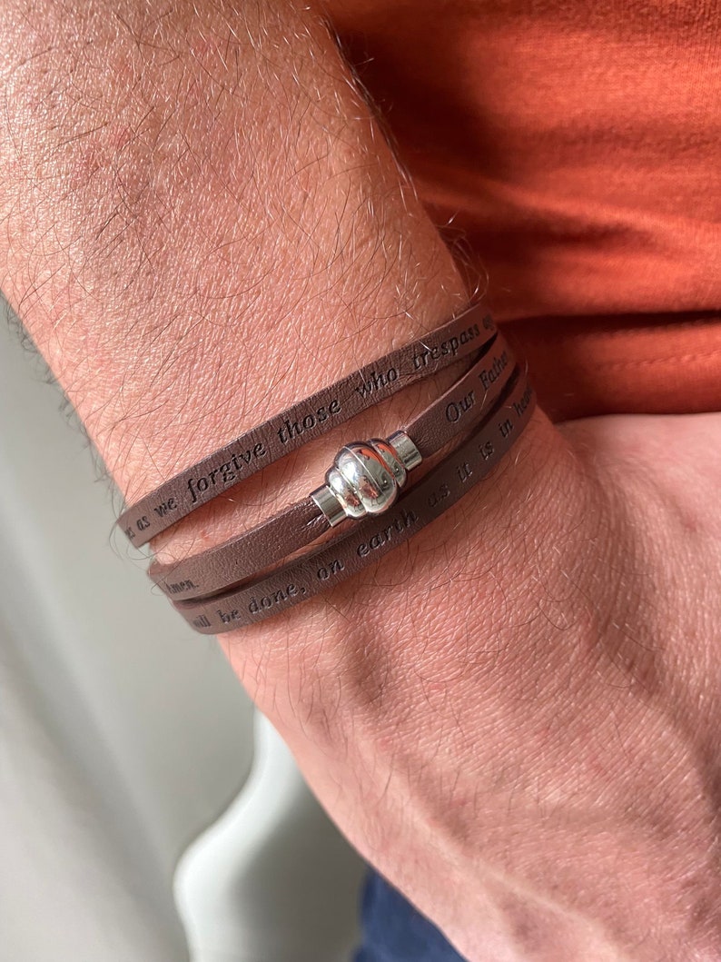 May include: A brown leather bracelet with a silver clasp. The bracelet has the words "as we forgive those who trespass against us" and "Our Father, who art in heaven, hallowed be thy name" engraved on it.