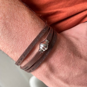 May include: A brown leather bracelet with a silver clasp. The bracelet has the words "Forgive us as we forgive those who trespass against us" engraved on it.