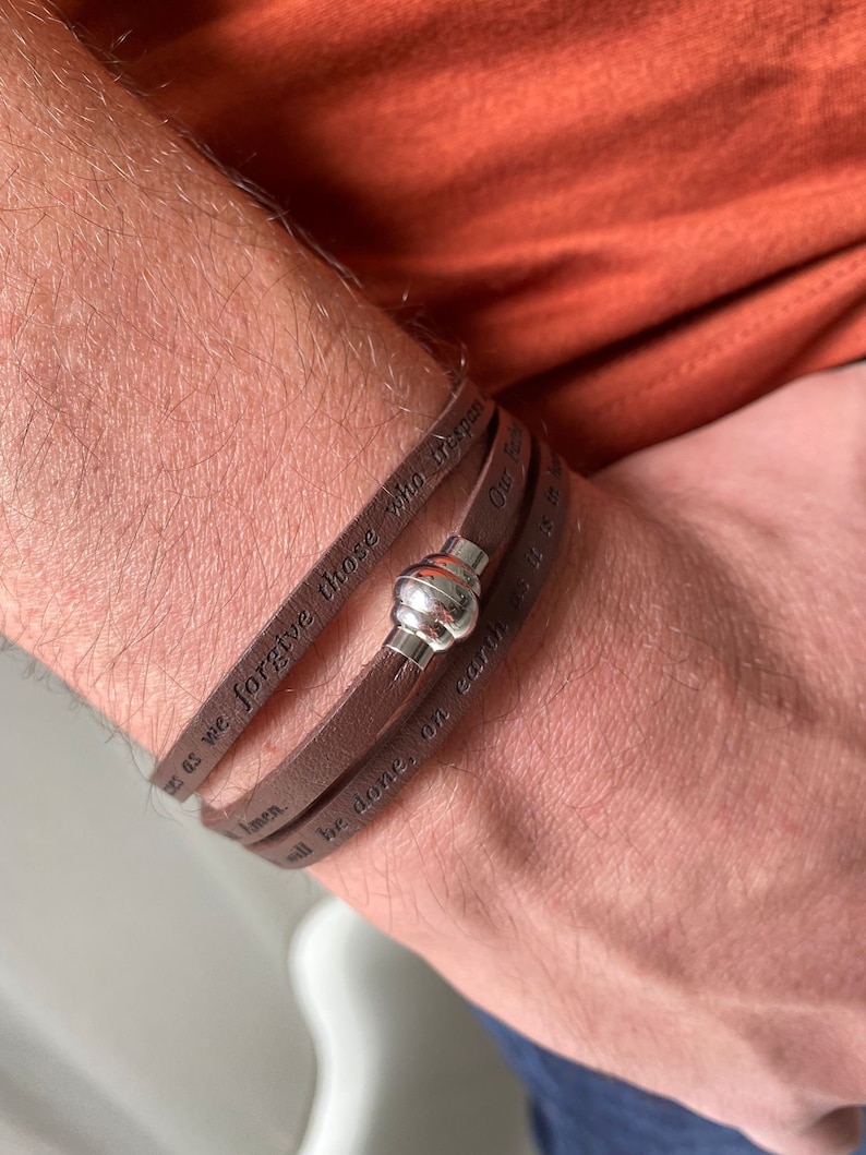 May include: A brown leather bracelet with a silver clasp. The bracelet has the words "Forgive us as we forgive those who trespass against us" engraved on it.