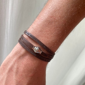 May include: A brown leather bracelet with a silver clasp. The bracelet has the words "Our Father" and "we forgive those who trespass against us" engraved on it.