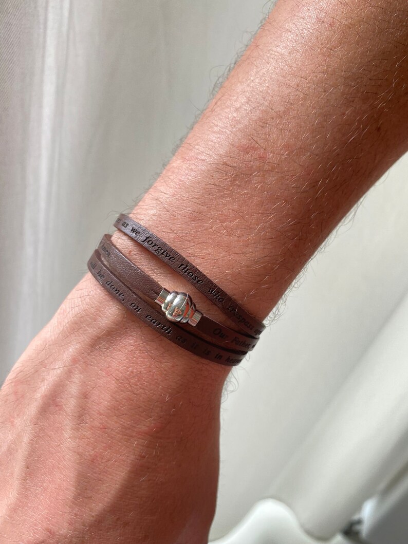 May include: A brown leather bracelet with a silver clasp. The bracelet has the words "Our Father" and "we forgive those who trespass against us" engraved on it.