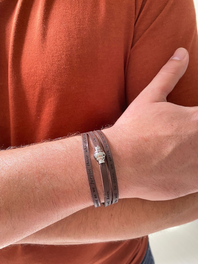 May include: A brown leather bracelet with a silver charm and the text "Forgive those who trespass against us as we forgive those who trespass against us. Amen. A will be done, on earth as it is in heaven."