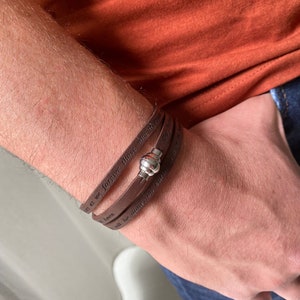 May include: Brown leather bracelet with a silver clasp. The bracelet has the words "Forgive those who trespass against us" and "All be done on earth as it is in heaven" engraved on it.