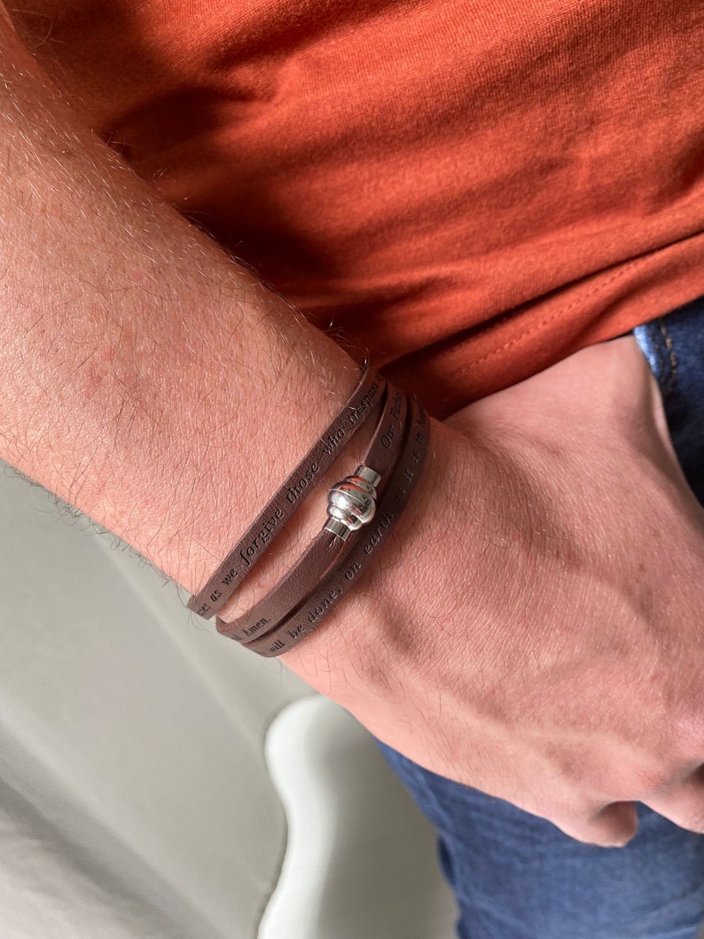 May include: Brown leather bracelet with a silver clasp. The bracelet has the words "Forgive those who trespass against us" and "All be done on earth as it is in heaven" engraved on it.