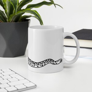Puede incluir: Taza de cerámica blanca con una ilustración en blanco y negro de una serpiente. El diseño de la serpiente envuelve la taza. La taza tiene un asa curva. El fondo incluye una planta y libros.
