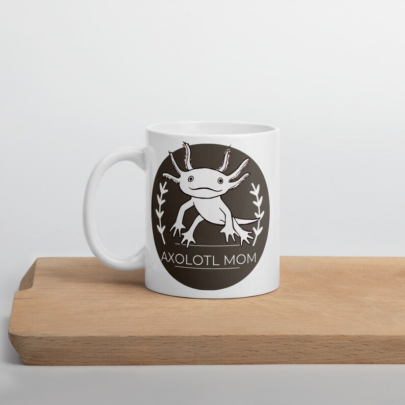 Puede incluir: Taza de cer&aacute;mica blanca con un dise&ntilde;o de ajolote en blanco y negro dentro de un c&iacute;rculo marr&oacute;n oscuro, con el texto "AXOLOTL MOM". La taza tiene un asa curva y est&aacute; colocada sobre una superficie de madera.