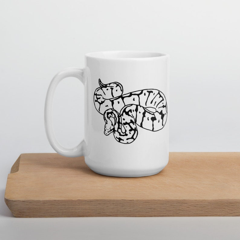 K&ouml;nnte beinhalten: Wei&szlig;e Keramiktasse mit einer Schwarz-Wei&szlig;-Illustration einer Schlange. Die Schlange hat einen detaillierten, gemusterten K&ouml;rper. Die Tasse hat einen gebogenen Griff und steht auf einer Holzoberfl&auml;che.