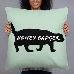 May include: A square, pale green cushion with a black honey badger silhouette. The words "HONEY BADGER" are printed in white across the badger's body. The cushion is held up by a person.