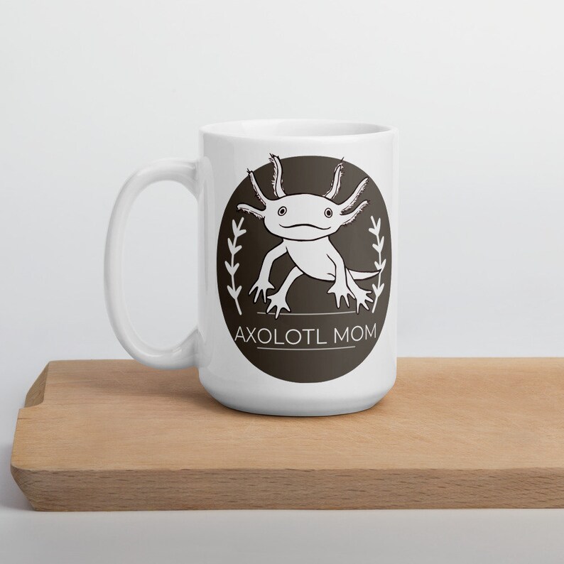 Puede incluir: Taza de cer&aacute;mica blanca con un dise&ntilde;o de ajolote en blanco y negro dentro de un c&iacute;rculo marr&oacute;n oscuro. El texto "AXOLOTL MOM" est&aacute; debajo. La taza est&aacute; sobre una tabla de cortar de madera.