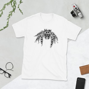 Puede incluir: Camiseta blanca con una ilustraci&oacute;n detallada en blanco y negro de una ara&ntilde;a saltarina. El dise&ntilde;o de la ara&ntilde;a est&aacute; centrado en la camiseta, mostrando sus patas peludas y sus ojos distintivos. La camiseta es de material suave.