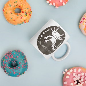 Puede incluir: Taza de cer&aacute;mica blanca con un dise&ntilde;o de ajolote en blanco y negro y el texto "AXOLOTL MOM". La taza est&aacute; rodeada de coloridos donuts con chispas sobre un fondo azul claro.