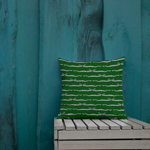 May include: A square decorative pillow with a green background and a repeating pattern of white and gray snakes. The pillow is resting on a white wooden crate against a teal wooden wall.