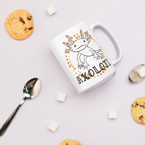 以下が含まれることがあります： 白のセラミックマグカップ。黒と白のアホロートルイラストと「AXOLOTL」の文字が描かれています。デザインはオレンジ色の点で囲まれています。マグカップは、クッキー、角砂糖、スプーンと一緒に、薄紫色の表面に置かれています。