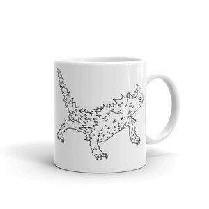 Pode incluir: Caneca de cerâmica branca com um desenho a traço preto de um lagarto diabo espinhoso. O lagarto é retratado em perfil, com espinhos detalhados e um corpo texturizado. A caneca tem uma alça padrão e um acabamento brilhante.