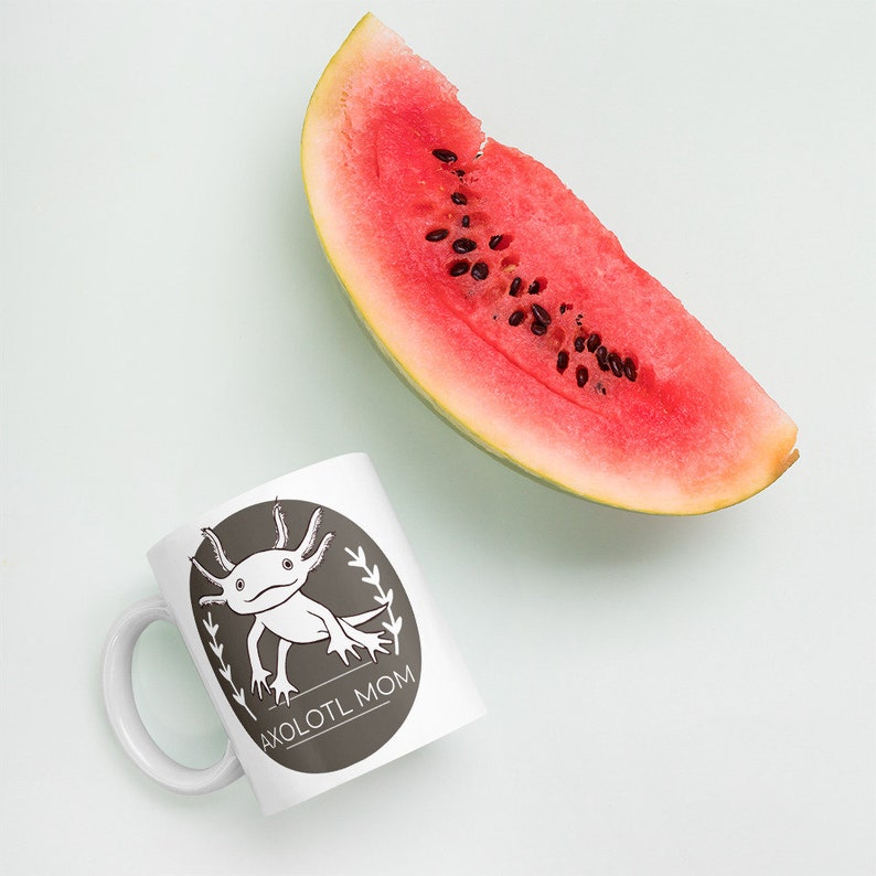 Puede incluir: Taza de cer&aacute;mica blanca con un dise&ntilde;o ovalado marr&oacute;n que presenta un ajolote de dibujos animados y el texto "AXOLOTL MOM". Una rodaja de sand&iacute;a est&aacute; al lado de la taza. La sand&iacute;a tiene un interior rojo y semillas negras.