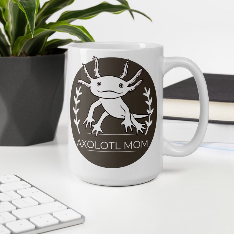 Puede incluir: Taza de cer&aacute;mica blanca con un dise&ntilde;o de ajolote de dibujos animados dentro de un &oacute;valo marr&oacute;n oscuro con el texto "AXOLOTL MOM". La taza tiene un asa curva y es adecuada para bebidas fr&iacute;as o calientes.