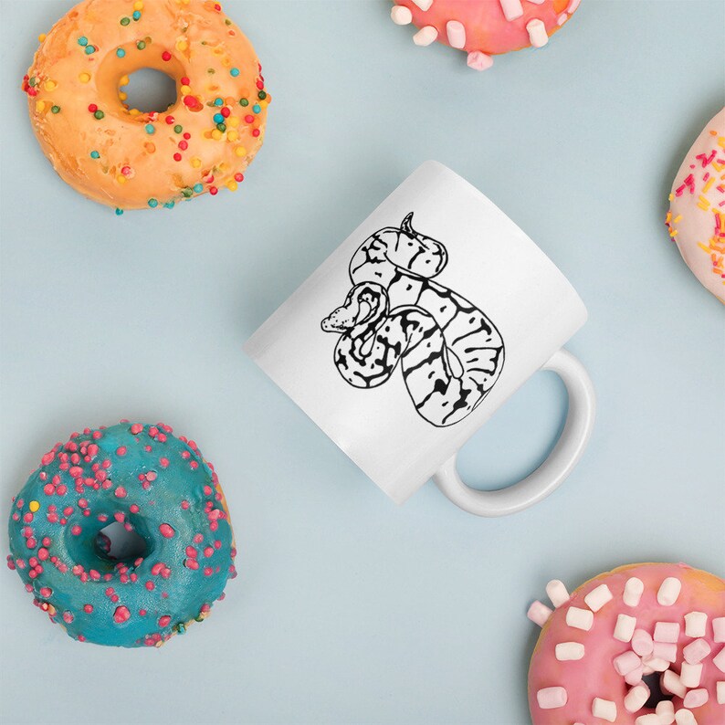 K&ouml;nnte beinhalten: Wei&szlig;e Keramiktasse mit einer schwarz-wei&szlig;en Illustration einer Schlange. Die Tasse ist von bunten Donuts mit Streuseln und Zuckerguss auf einer hellblauen Oberfl&auml;che umgeben.