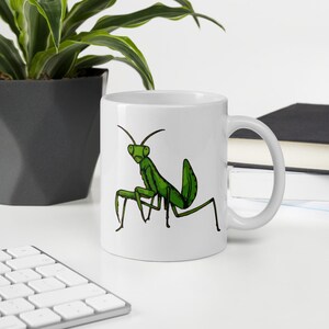 May include: White ceramic mug featuring a green cartoon praying mantis illustration. The insect is depicted with a detailed outline and a vibrant green color. The mug has a standard handle and is suitable for hot or cold beverages.