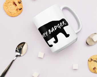 May include: White ceramic mug with a black silhouette of a honey badger and the words "HONEY BADGER" in white. The mug is surrounded by cookies, sugar cubes, and a spoon.