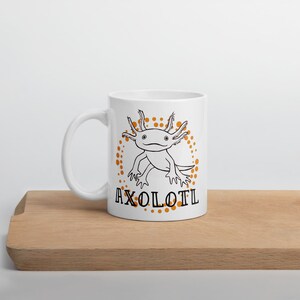 以下が含まれることがあります： 白のセラミックマグカップ。黒と白のアホロートルイラストと「AXOLOTL」の文字が特徴です。アホロートルはオレンジ色の点で囲まれています。マグカップは湾曲したハンドルがあり、木製の表面に置かれています。