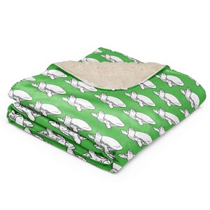 Könnte beinhalten: Eine gefaltete grüne Decke mit einem weißen Schildkrötenmuster. Die Decke hat ein weiches, cremefarbenes Sherpa-Futter. Das Schildkrötenmuster wiederholt sich über die grüne Oberfläche.