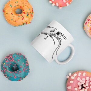 Op de afbeelding: Witte keramische mok met een zwarte lijntekening van een garnaal. De mok is omringd door kleurrijke donuts met hagelslag op een lichtblauwe achtergrond. De donuts hebben oranje, roze en blauwe glazuur.