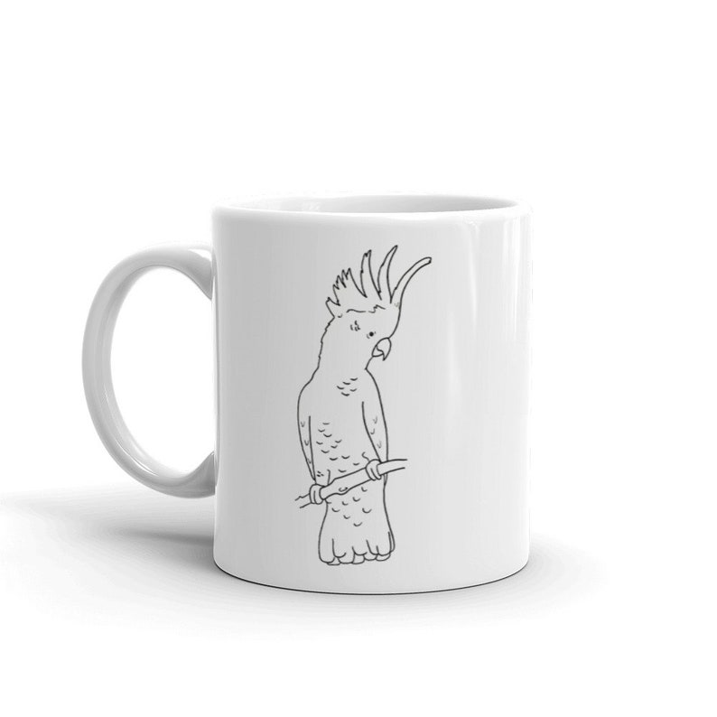 May include: White ceramic mug featuring a black line drawing of a cockatoo perched on a branch. The bird has a distinctive crest and is facing to the right. The mug has a curved handle and a glossy finish.
