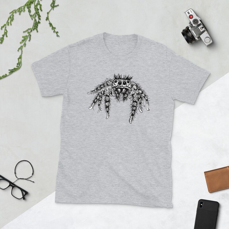 Puede incluir: Una camiseta gris claro con una ilustraci&oacute;n detallada en blanco y negro de una ara&ntilde;a saltarina. La ara&ntilde;a tiene ojos grandes y patas peludas. La camiseta est&aacute; hecha de material suave y es adecuada para uso casual.