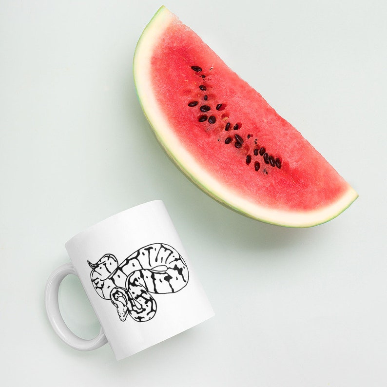 K&ouml;nnte beinhalten: Wei&szlig;e Keramiktasse mit einer schwarz-wei&szlig;en Illustration einer Schlange. Eine gro&szlig;e Scheibe Wassermelone mit rotem Fruchtfleisch und schwarzen Samen liegt neben der Tasse auf hellblauem Hintergrund.