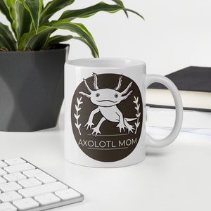 Puede incluir: Taza de cer&aacute;mica blanca con un dise&ntilde;o de ajolote de dibujos animados dentro de un c&iacute;rculo marr&oacute;n oscuro, el texto dice "AXOLOTL MOM". La taza tiene un asa curva. Una planta y un teclado est&aacute;n en el fondo. La taza est&aacute; sobre una superficie blanca.