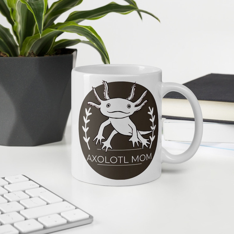 Puede incluir: Taza de cer&aacute;mica blanca con un dise&ntilde;o de ajolote de dibujos animados dentro de un c&iacute;rculo marr&oacute;n oscuro, el texto dice "AXOLOTL MOM". La taza tiene un asa curva. Una planta y un teclado est&aacute;n en el fondo. La taza est&aacute; sobre una superficie blanca.